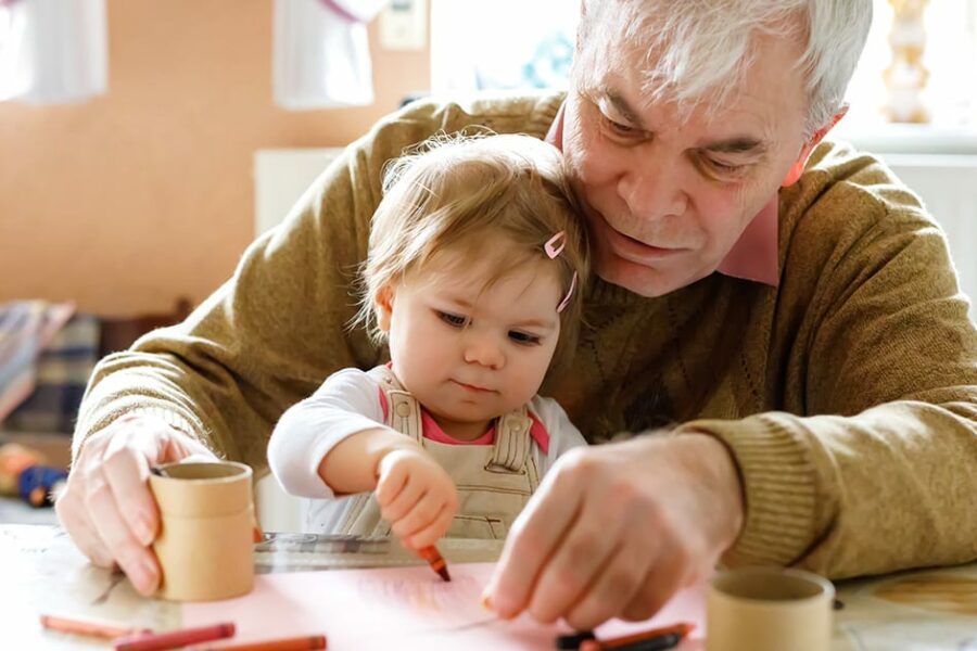 Cute little baby toddler girl and handsome senior grandfather painting with colorful pencils at home. Grandchild and man having fun together